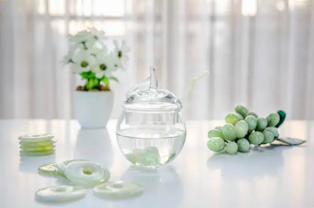 Polished small jade stones are located inside of a glass jar with crystalline water. It is making crystal charged water at home. There are donuts carved grapes and white flowers on the table.