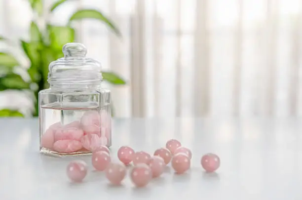 Tumbled rose quartz stones are placed inside of a glass jar with drinking water. It is a gem elixir preparation. Several beads lie in the foreground out of focus.