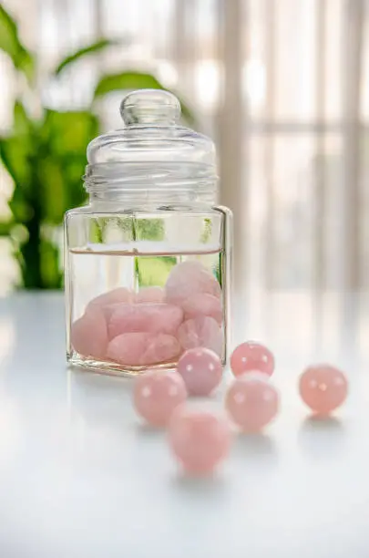Burnished rose quartz stones are put inside of a glass jar. The drinking water is being charged with the healing energy of crystals. Six  beads are in the foreground out of focus.