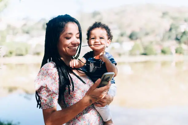 Young African descendant mother her baby son outdoors using smartphone