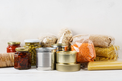 Various canned food and raw cereal grains on a table. Set of grocery goods for cooking, delivery or donation. Copy space.