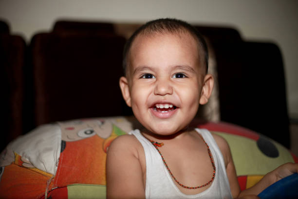Portrait Of Smiling Healthy Baby In White Bodysuit Sitting On Bed. Copy Space
