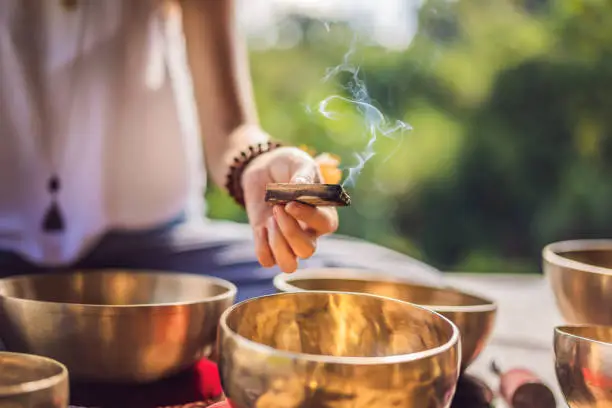 Photo of Woman playing on Tibetan singing bowl while sitting on yoga mat against a waterfall. Vintage tonned. Beautiful girl with mala beads meditating