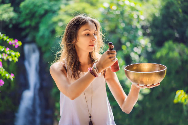 femme jouant sur le bol de chant tibétain tout en s'asseyant sur le tapis de yoga contre une chute d'eau. vintage tonned. belle fille avec des perles de mala méditant - tibetan culture music sound recovery photos et images de collection