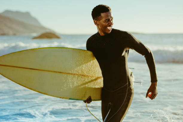 Smiling african male surfer Smiling african male running out of the ocean after water surfing. Happy young man with surfboard on the beach enjoying holidays. wetsuit stock pictures, royalty-free photos & images