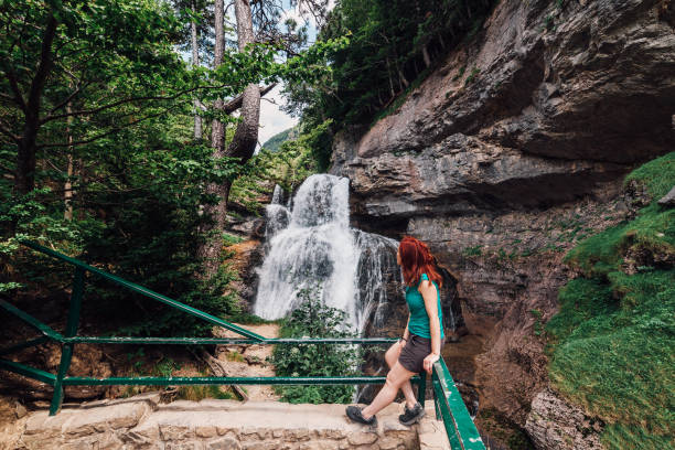 istock Young woman looking at a waterfall in the forest among trees 1162960040