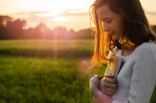 Christian teenage girl holds bible in her hands. Reading the Holy Bible in a field during beautiful sunset. Concept for faith, spirituality and religion
