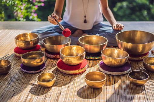 Woman playing on Tibetan singing bowl while sitting on yoga mat against a waterfall. Vintage tonned. Beautiful girl with mala beads meditating.