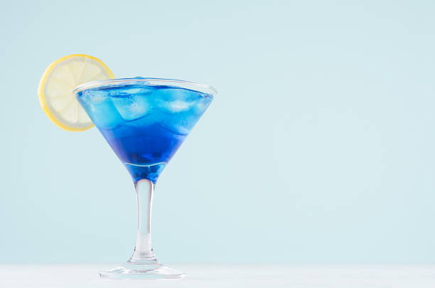istock Tropical fresh alcohol cocktail with blue curacao liqueur, ice cube, lemon slice in misted martini glass on soft light white, pastel green background. 1162964316