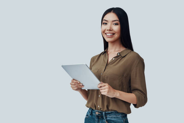 istock Technologies in her hands. Attractive young Asian woman looking at camera and smiling while standing against grey background 1162966186