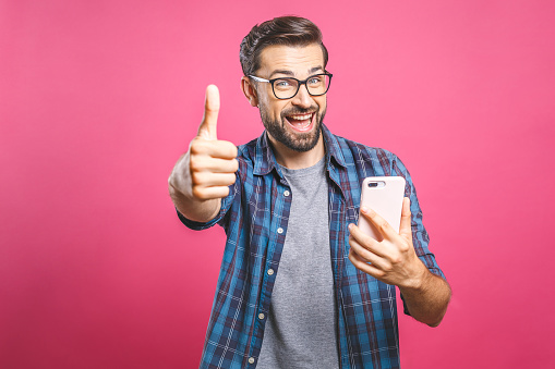 Portrait of a cheerful bearded man taking selfie and showing thumbs up gesture over pink background. Isolated.