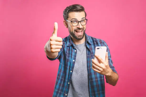 Portrait of a cheerful bearded man taking selfie and showing thumbs up gesture over pink background. Isolated.