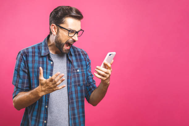 Young caucasian man angry, frustrated and furious with his phone, angry with customer service over pink background. Young caucasian man angry, frustrated and furious with his phone, angry with customer service over pink background. displeased stock pictures, royalty-free photos & images