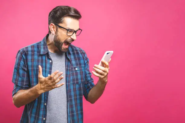 Photo of Young caucasian man angry, frustrated and furious with his phone, angry with customer service over pink background.