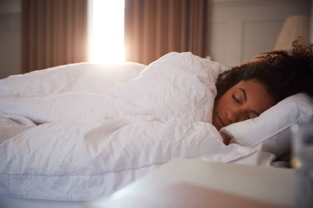 peaceful woman asleep in bed as day break through curtains - deitando imagens e fotografias de stock