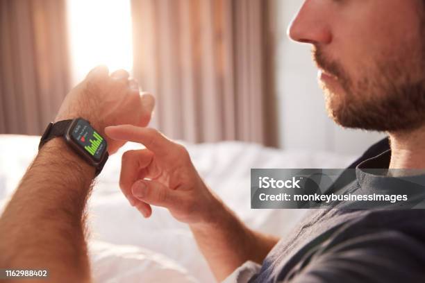 Man Sitting Up In Bed Looking At Screen Of Smart Watch Stock Photo - Download Image Now