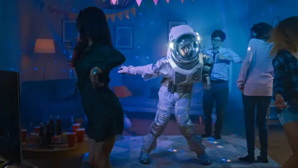 Photo of At the College House Costume Party: Fun Guy Wearing Space Suit Dances Off, Doing Robot Dance Modern Moves. With Him Beautiful Girls and Boys Dancing in Neon Lights.