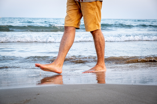 Legs of a man with short trousers walking along the shore of a mediterranean beach at sunset
