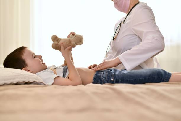 The doctor examines the baby tummy with a bear. Doctor on call at home of a child who has a stomach ache. A contented boy is lying on the bed holding a teddy bear in his hands. bear stomach stock pictures, royalty-free photos & images