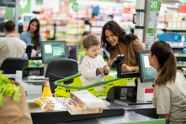 madre amorosa enseñando a su hijo donde poner la tarjeta de crédito para pagar los comestibles en la caja mientras está de pie en el carro de la compra todo sonriendo - checkout counter cash register retail supermarket fotografías e imágenes de stock