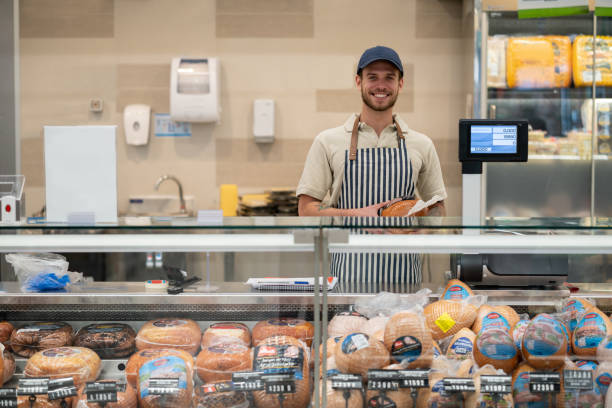 retrato de joven vendedor de pie detrás del mostrador de la sección delicatessen frente a la cámara muy feliz - delicatessen fotografías e imágenes de stock