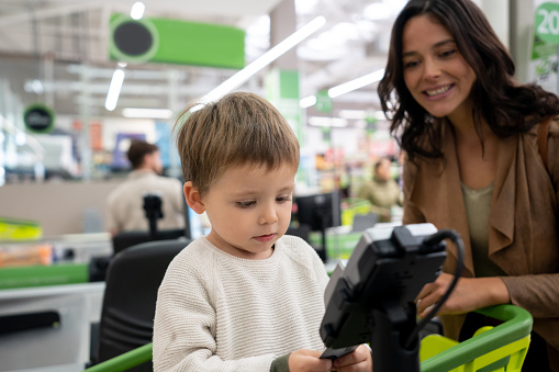 Cute baby boy introducing card on credit card reader at checkout of the supermarket while mom supervises very cheerfully and smiling