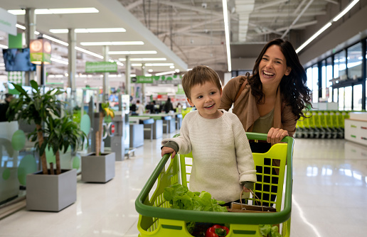 Hermosa madre empujando el carro con su niño pequeño en el interior con comestibles comprados salir del supermercado y frente a la cámara sonriendo photo