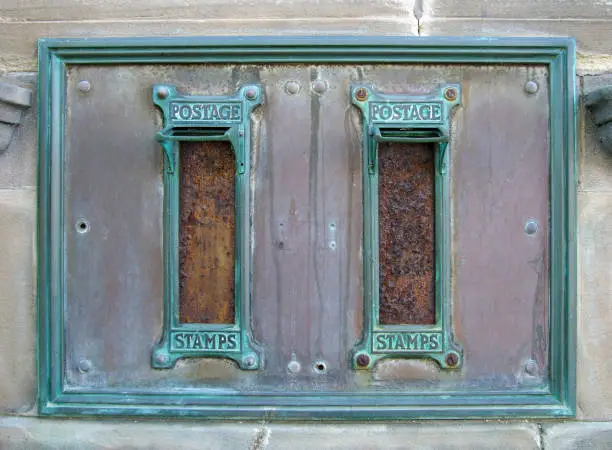 Photo of old british postal mail boxes with rusted letter slots and ornate green copper frames with the words postage stamps surrounded by an old stained metal frame in a stone wall