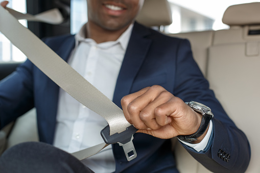 Young african american businessman sitting on the back seat in the car fastening seat belt close-up blurred background smiling cheerful