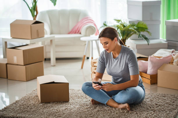 istock Woman using phone while sitting on living room floor, surrounded by boxes 1163060573