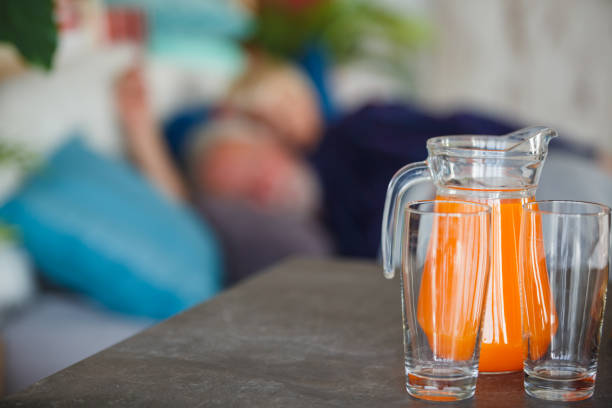 Copy space shot of two empty glasses next to a pitcher with orange juice laid on a kitchen counter while in the blurred background senior married couple is sleeping in their bed.