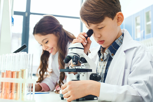 Little boy and girl learning in school laboratory boy looking in a microscope concentrated making experiment girl writing results smiling happy