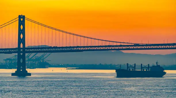 Foggy/Misty summer morning sunrise in California and silhouette of the iconic Oakland Bay Bridge in San Francisco as cargo ship sails by. Famous travel location landmark in the west coast city.