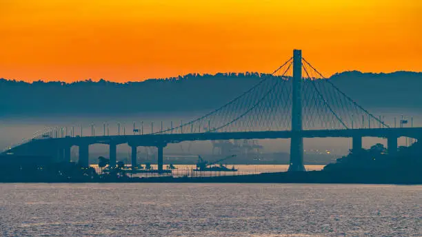 Beautiful summer morning sunrise behind mountain in California gives silhouette to the iconic Oakland Bay Bridge in San Francisco over the ocean. Famous travel location landmark in the west coast city.