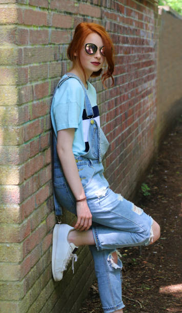 Stock photo of pretty moody, sad teenage girl youth with red hair looking at camera, smiling lost in thought by brick wall, redhead wearing blue denim dungarees, sunglasses, makeup, dark red lipstick, teenager with ginger hair tied back in curls imagen de bastante malhumorado, triste adolescente adolescente con el pelo rojo mirando a la cámara, sonriendo perdido en el pensamiento por la pared de ladrillo, pelirroja usando dungarees de mezclilla azul, gafas de sol, maquillaje, lápiz labial rojo o