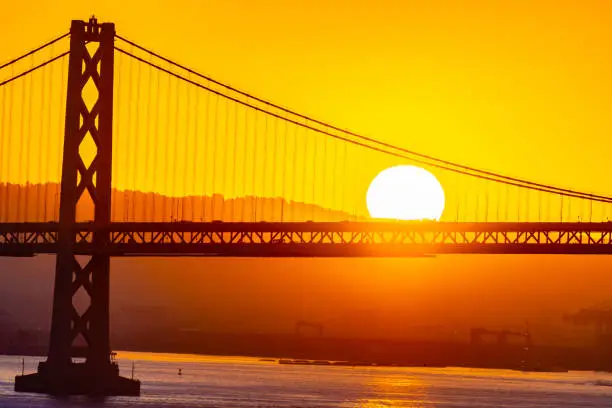 Beautiful summer morning sunrise behind mountain in California gives silhouette to the iconic Oakland Bay Bridge in San Francisco over the ocean. Famous travel location landmark in the west coast city.