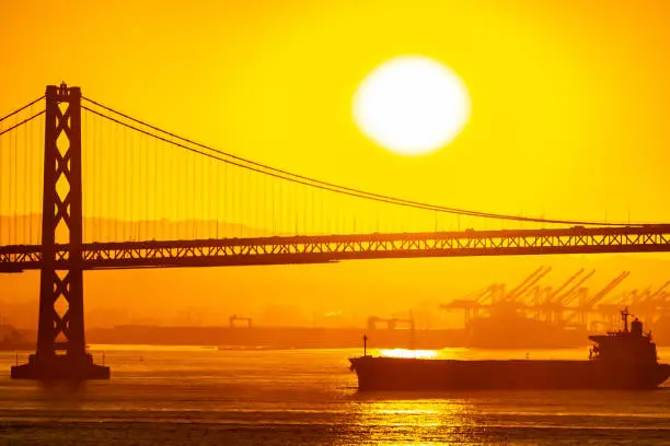 Foggy/Misty summer morning sunrise in California and silhouette of the iconic Oakland Bay Bridge in San Francisco as cargo ship sails by. Famous travel location landmark in the west coast city.