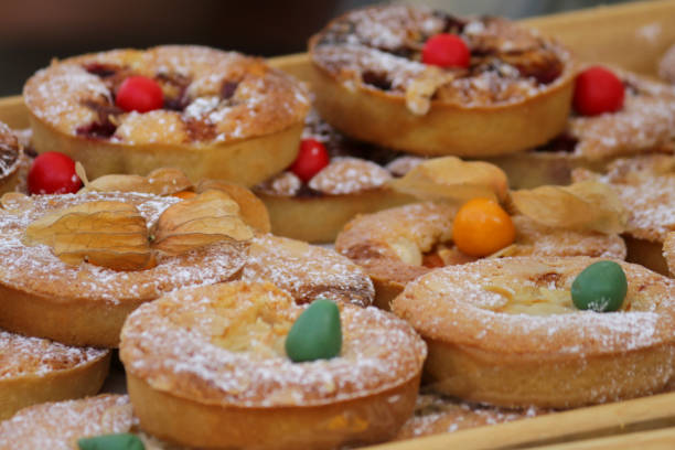 Stock photo of delicious sweet homemade cherry bakewell tarts, pies and pasties for sale at street food market bakery stall, topped with Chinese lanterns fruit (Physalis Pubescens) and dusted with icing sugar, ready to eat for unhealthy diet afternoon snack imagen de deliciosas tartas dulces caseras de pastel de cerezo, pasteles y pasteles a la venta en el puesto de panadería del mercado de comida callejera, cubierto con fruta de linterna china (physalis pubescens) y espolvoreado con azúcar glas, listo para