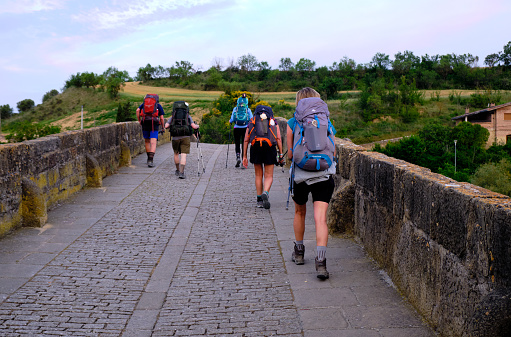 Navarre, Spain, June 3 2019: Pilgrims walking on the Camino de Santiago.  Seen from back Leaving early in morning by crossing the historical bridge in Puente la Reina