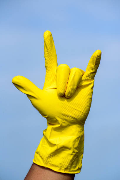 Image of Human hand in yellow protective rubber glove showing Rock On sign with blue sky background.