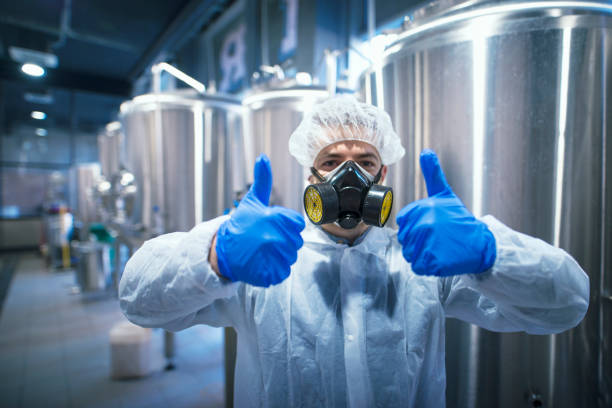 istock Professional technologist expert in protective white uniform with hairnet, mask and gloves holding both thumbs up. Industrial worker specialist celebrating good results in food processing industry. 1163142874