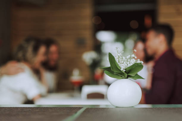 Close up of table with lilies of the valley in round white vase. Young people spending time in cafe