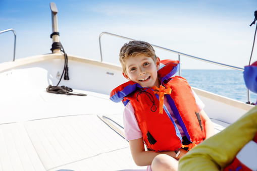 Happy blonde kid boy enjoying sailing boat trip. Family vacations on ocean or sea on sunny day. Healthy beautiful school child smiling and having fun on yacht. Coastline with villages and nature.