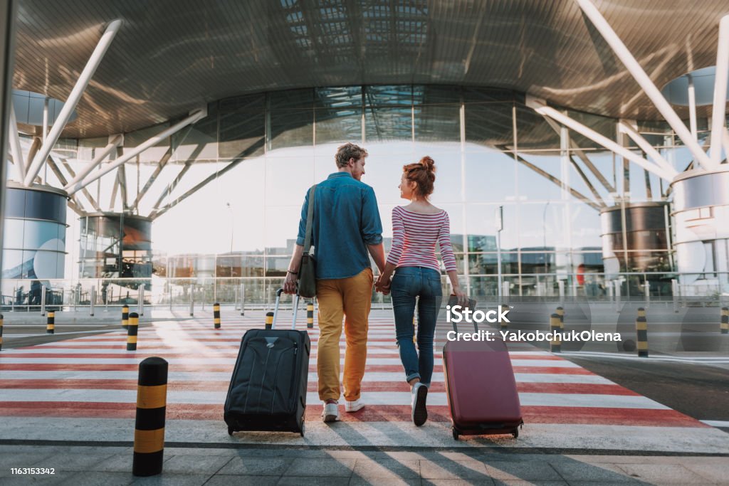 Beautiful loving couple with travel suitcases holding hands in airport Full length back view portrait of young man and his charming girlfriend walking and carrying their trolley bags. They looking at each other and smiling Airport Stock Photo