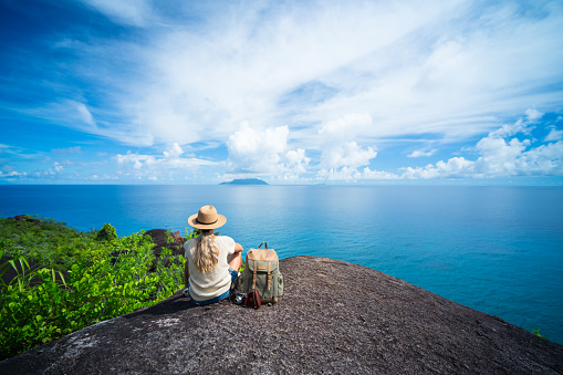 rear view woman with backpack, old manual photo camera and straw hat  sitting relaxing on granite rock watching the calm indian ocean on sunny vacation day