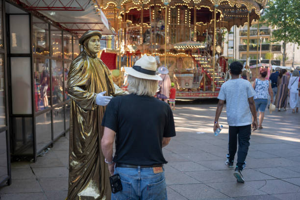 The Avignon Festival is an annual arts festival which takes place in Avignon, in the south of France. On July 12, 2019 during this festival, in Avignon, France a tourist chats with a performer draped in gold. In the background is the two-level La Belle Epoque carousel.