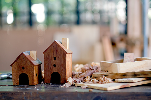 Two small wood houses or bird box are put on the table among wood pile and other equipment for crafting in the room with soft light.