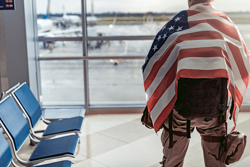 Back view of adult man wearing military uniform and situating in waiting room while carrying his backpack. He is covering with American flag while standing opposite the window. Serving the state concept