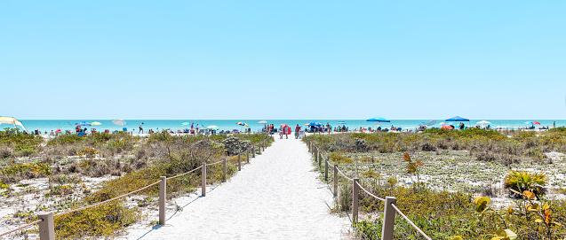 Panorama of Bowman's beach at Sanibel Island with sandy trail, path, walkway, fence, many people, crowd in distance, crowded coast, coastline shelling, looking for shells