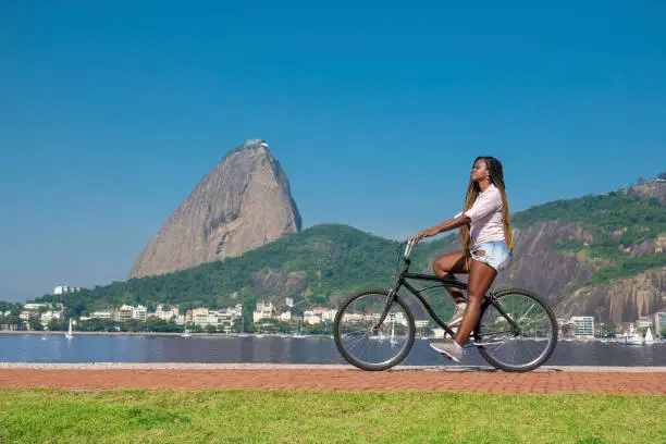 Beautiful black brazilian woman rides on the bike path in front of the Sugarloaf in Flamengo Park